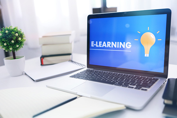 Open computer featuting “E-learning” graphic on the screen sitting on a desk with books, notebooks and a small plant in front of a window with white curtains