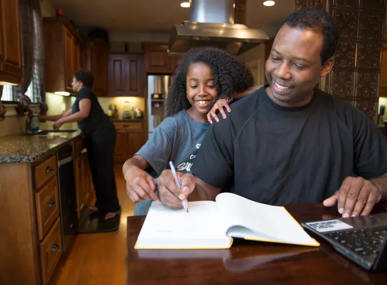 Black man studying at table with laptop and book with daughter standing behind him and wife at kitchen sink in background