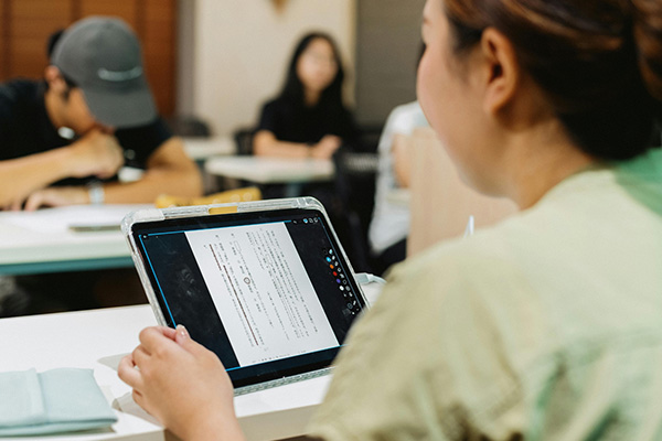 Over the shoulder closeup of a female student sitting in a classroom working on a tablet with other students out of focus in the distance