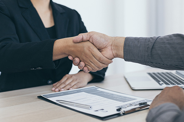 Unidentifiable dark skinned male and female sitting at a table or desk shaking hands with a clipboard and open laptop on the desk